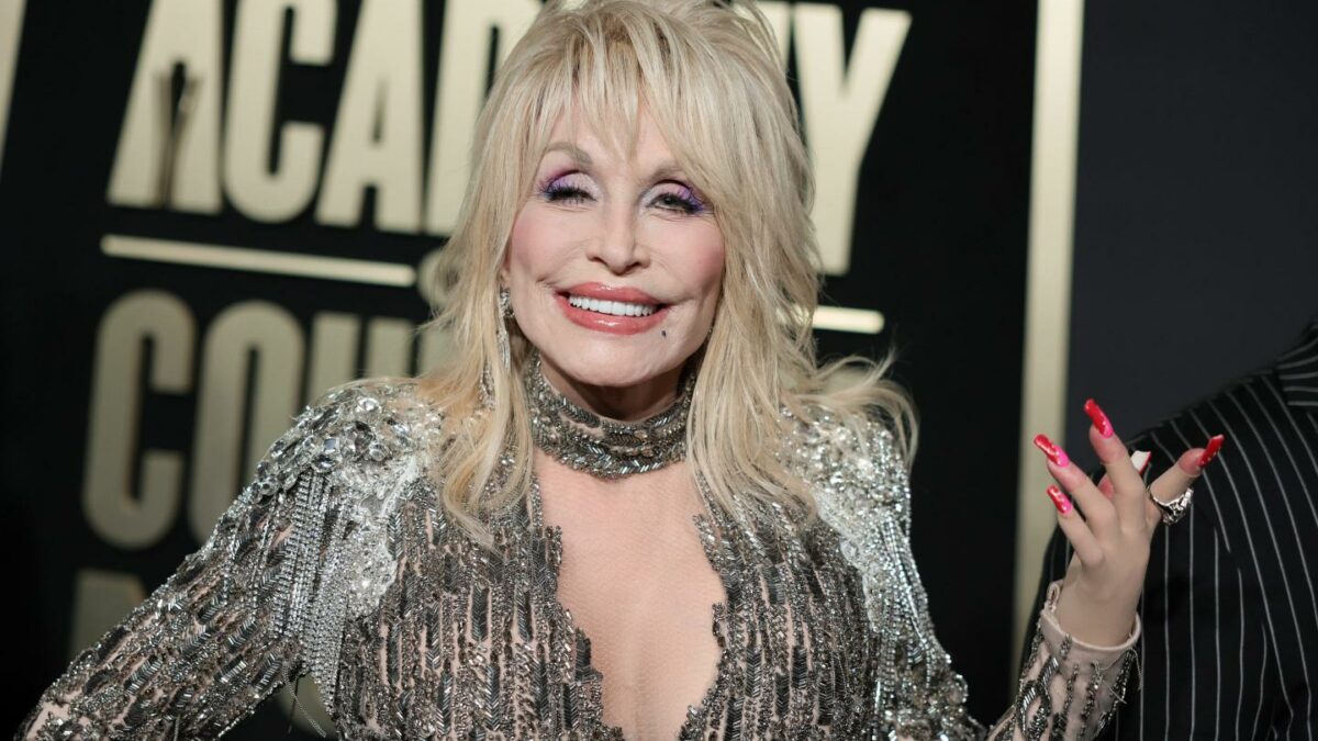 Dolly Parton smiles at the camera with her hand up and fingers spread apart.