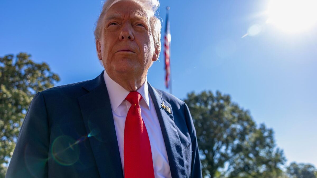 Trump in a suit and red tie with an American flag behind him and a backdrop of trees