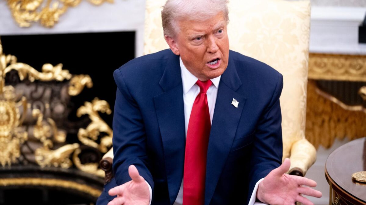 Trump sits in a chair in the Oval Office, speaking while gesturing with both hands.