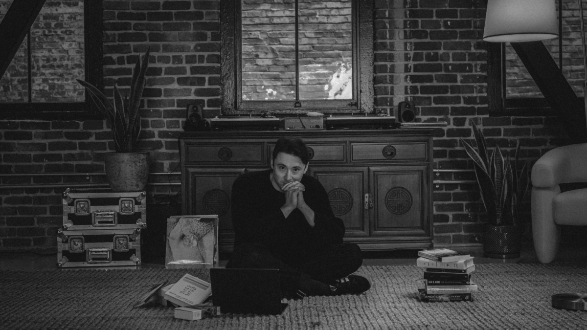Man sitting cross-legged on the floor of a carpeted room. There are stacks of books to either side of him.