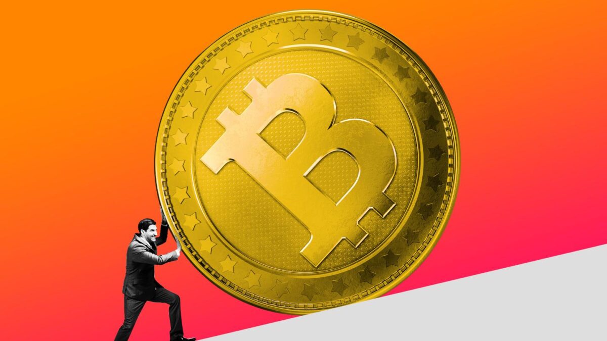 A businessman attempts to push a large stone bitcoin up a hill.